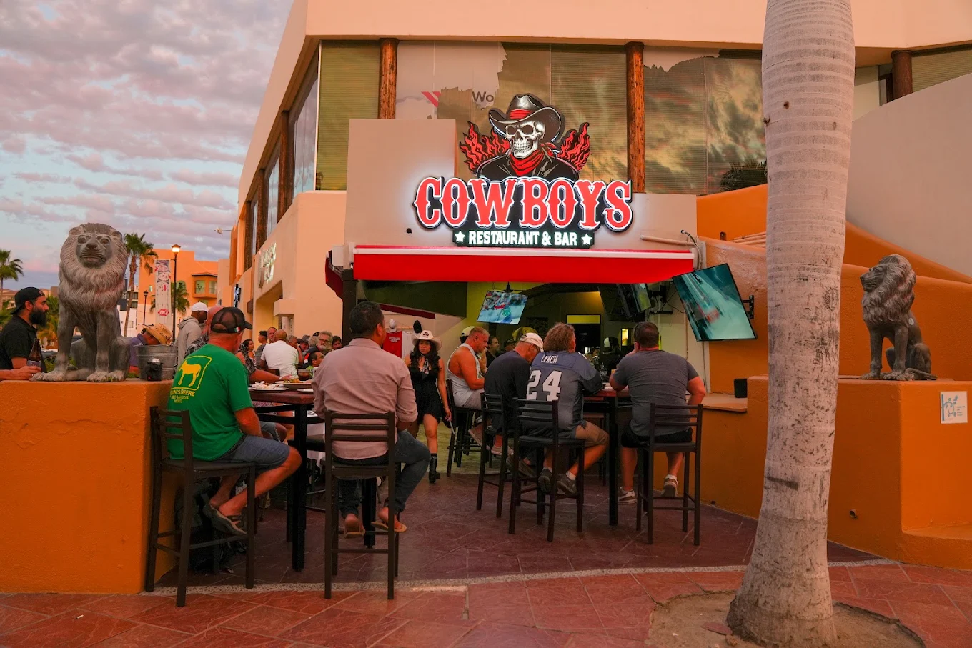 Crowd gathered on the covered patio under the glowing Cowboys Restaurant & Bar sign at dusk
